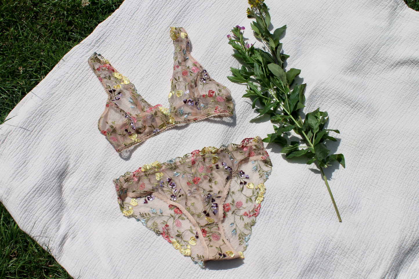 Embroidered flower bralette and knickers laid flat on a white picnic blanket with grass and flowers in the background